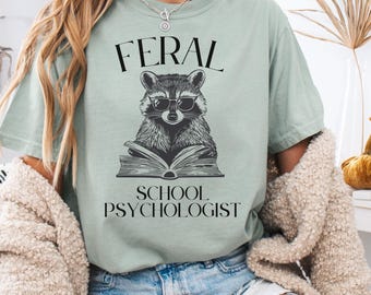 May include: Sage green t-shirt with a raccoon wearing sunglasses and reading a book. The text "FERAL SCHOOL PSYCHOLOGIST" is printed above and below the raccoon illustration. The shirt has short sleeves.
