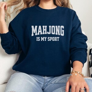 May include: Navy blue sweatshirt with the text "MAHJONG IS MY SPORT" in white block letters. The sweatshirt is a long-sleeved crew neck style. The person in the image is wearing blue jeans and a gold bracelet.