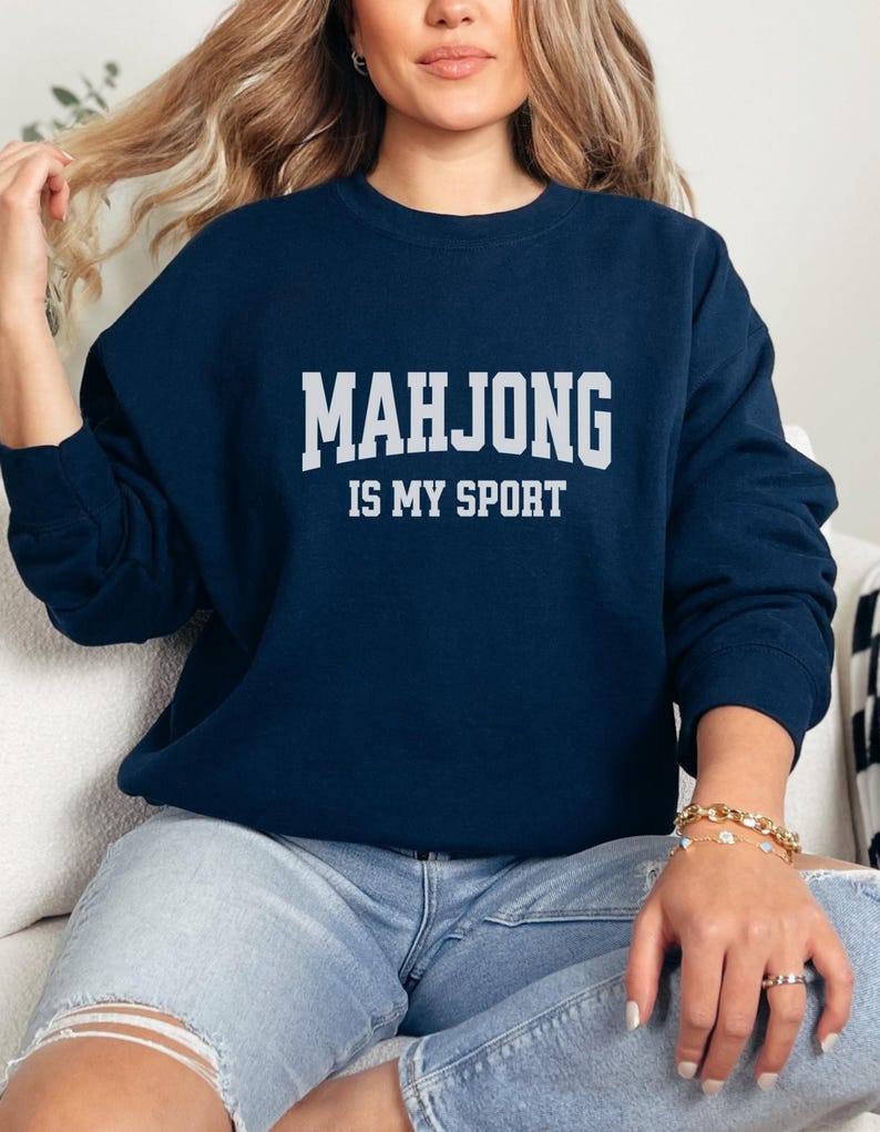 May include: Navy blue sweatshirt with the text "MAHJONG IS MY SPORT" in white block letters. The sweatshirt is a long-sleeved crew neck style. The person in the image is wearing blue jeans and a gold bracelet.