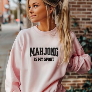 May include: A pale pink sweatshirt featuring the text "MAHJONG IS MY SPORT" in black. The crew neck sweatshirt has long sleeves. The image shows a person wearing the sweatshirt.