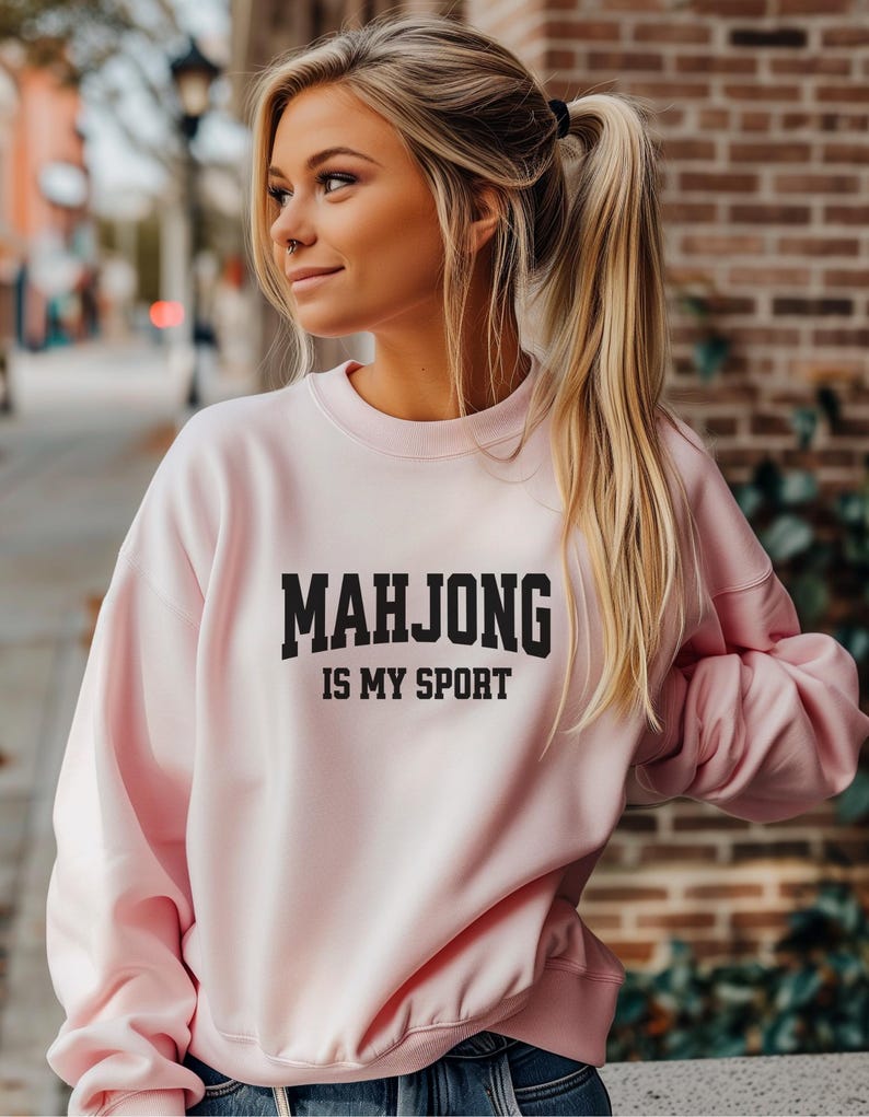 May include: A pale pink sweatshirt featuring the text "MAHJONG IS MY SPORT" in black. The crew neck sweatshirt has long sleeves. The image shows a person wearing the sweatshirt.