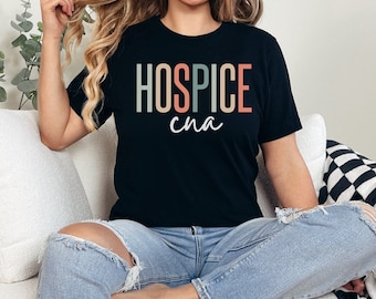Könnte beinhalten: Ein schwarzes T-Shirt mit dem Text "HOSPICE cna" in bunten Buchstaben. Das Shirt wird von einer Person getragen, die auf einem weißen Sofa sitzt.