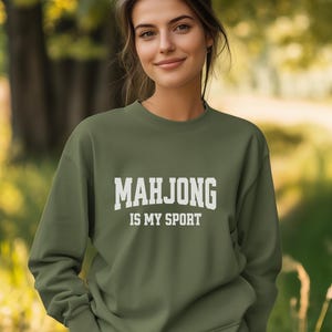 May include: An olive green sweatshirt with the text "MAHJONG IS MY SPORT" in bold white letters. The sweatshirt is a crew neck style and is being worn outdoors.