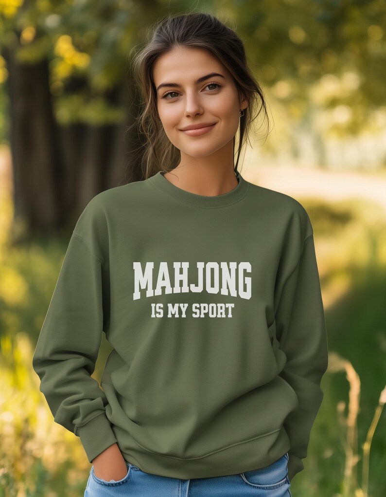 May include: An olive green sweatshirt with the text "MAHJONG IS MY SPORT" in bold white letters. The sweatshirt is a crew neck style and is being worn outdoors.