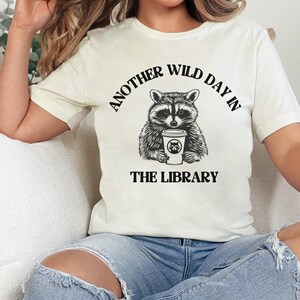 Peut inclure: T-shirt cr&egrave;me avec un dessin noir et blanc d'un raton laveur tenant une tasse de caf&eacute;. Le texte "ANOTHER WILD DAY IN THE LIBRARY" est arqu&eacute; au-dessus et en dessous de l'illustration du raton laveur. Le t-shirt a des manches courtes.
