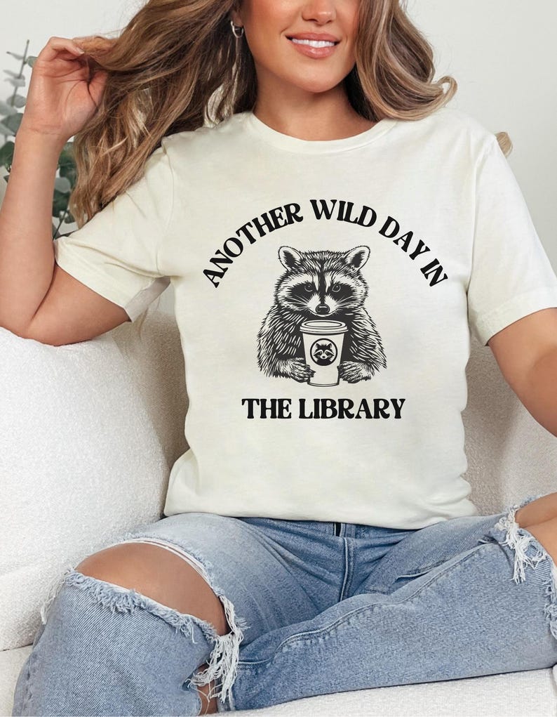 Peut inclure: T-shirt cr&egrave;me avec un dessin noir et blanc d'un raton laveur tenant une tasse de caf&eacute;. Le texte "ANOTHER WILD DAY IN THE LIBRARY" est arqu&eacute; au-dessus et en dessous de l'illustration du raton laveur. Le t-shirt a des manches courtes.