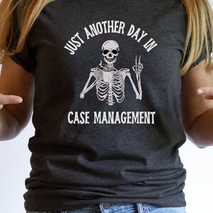May include: A dark gray t-shirt with a white graphic of a skeleton holding a coffee cup and making a peace sign. The text on the shirt reads "Just another day in case management."