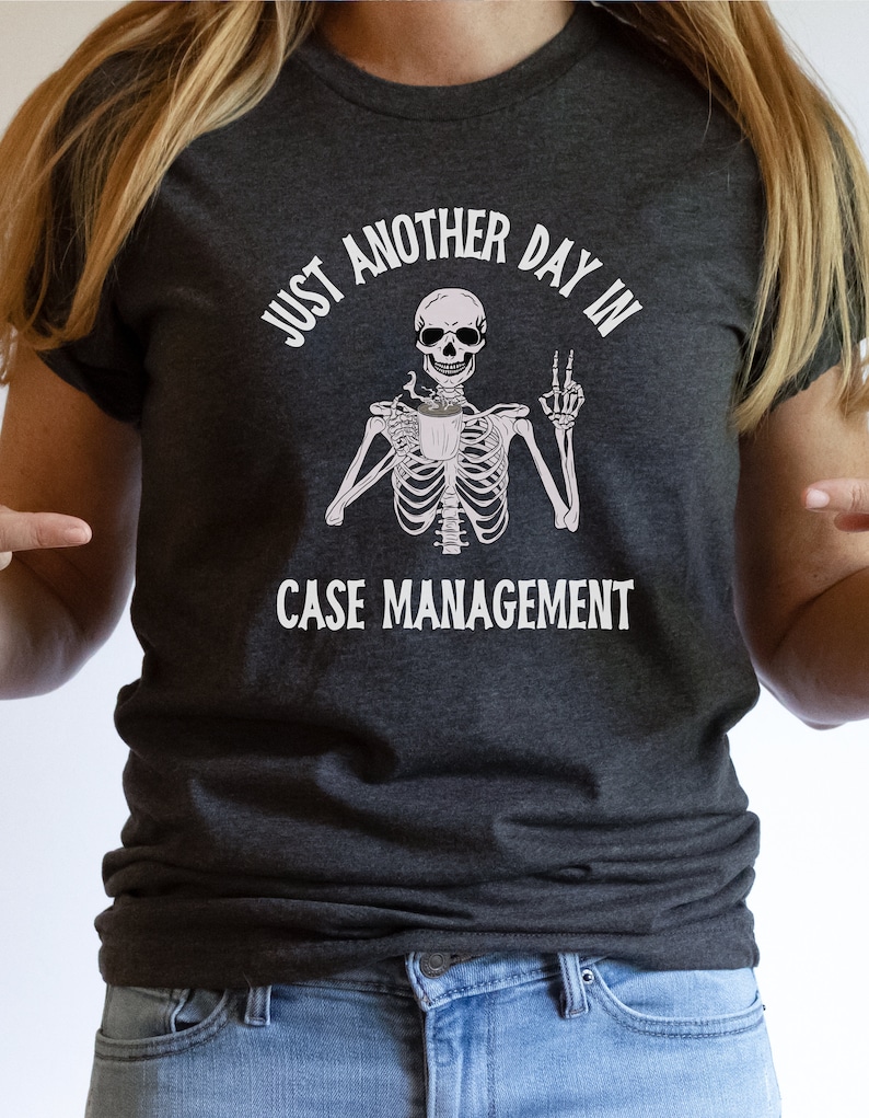 May include: A dark gray t-shirt with a white graphic of a skeleton holding a coffee cup and making a peace sign. The text on the shirt reads "Just another day in case management."