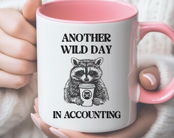 May include: White ceramic mug with a pink interior and handle. The mug features the text "ANOTHER WILD DAY IN ACCOUNTING" above and below a black and white illustration of a raccoon holding a coffee cup.