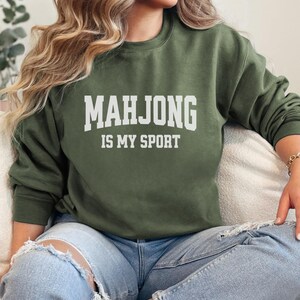 May include: An olive green sweatshirt with the text "MAHJONG IS MY SPORT" in white block letters. The sweatshirt is a crew neck style and is made of a soft material. The person is wearing ripped blue jeans.