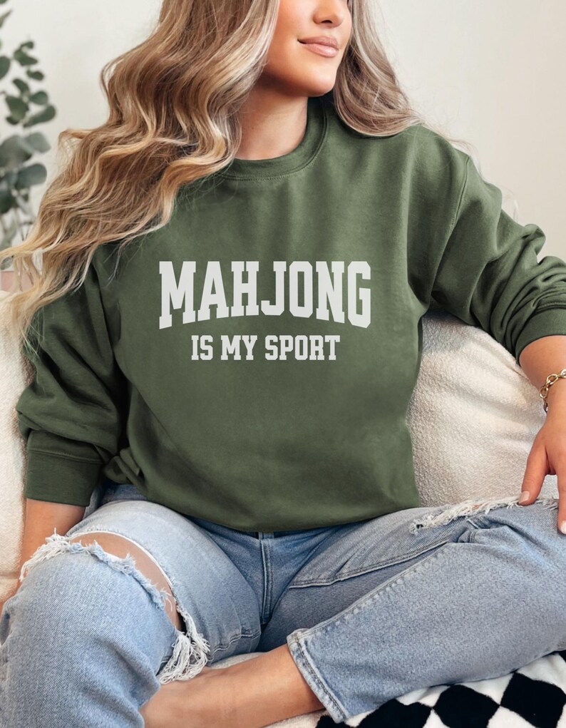 May include: An olive green sweatshirt with the text "MAHJONG IS MY SPORT" in white block letters. The sweatshirt is a crew neck style and is made of a soft material. The person is wearing ripped blue jeans.