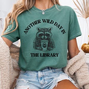 Peut inclure: T-shirt vert avec le texte "ANOTHER WILD DAY IN THE LIBRARY" au-dessus et en dessous d'un dessin de raton laveur tenant une tasse de caf&eacute;. Le t-shirt est de couleur verte unie.
