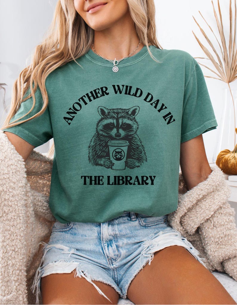 Peut inclure: T-shirt vert avec le texte "ANOTHER WILD DAY IN THE LIBRARY" au-dessus et en dessous d'un dessin de raton laveur tenant une tasse de caf&eacute;. Le t-shirt est de couleur verte unie.