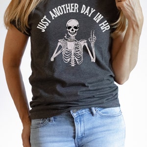 May include: A dark gray t-shirt with a skeleton holding a coffee cup and the text "Just another day in HR".