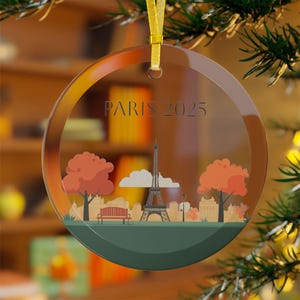 May include: A clear glass ornament with a gold ribbon and a printed image of the Eiffel Tower, trees, and the text "PARIS 2025".
