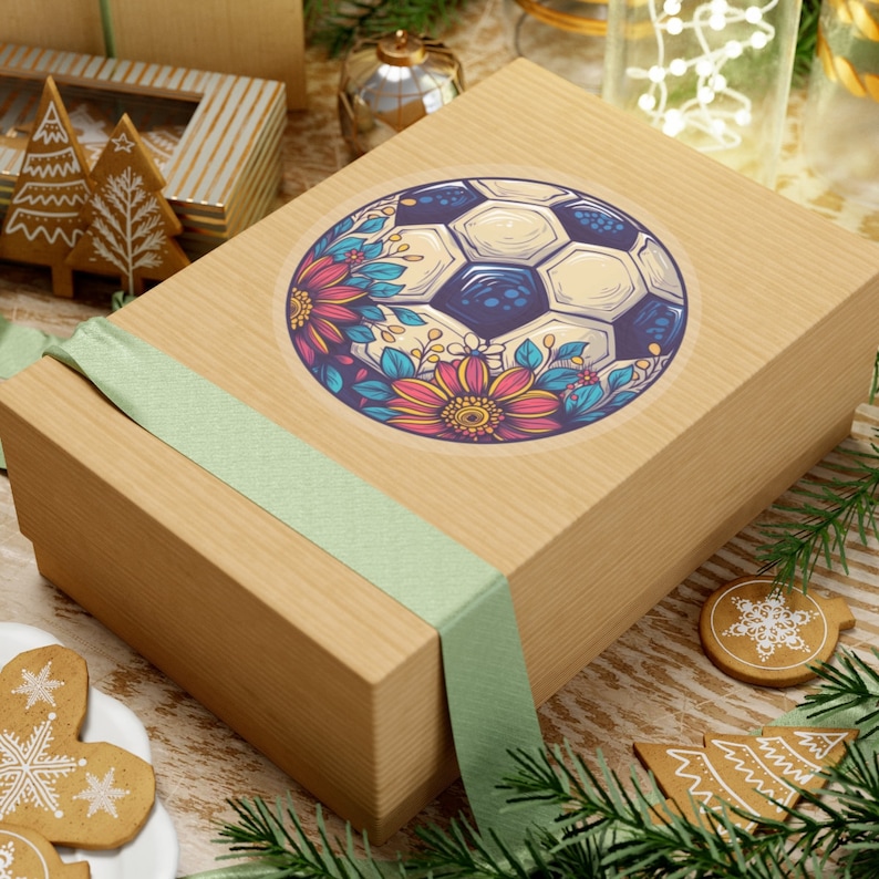 Puede incluir: Una caja de regalo marr&oacute;n con una cinta verde atada alrededor. La caja tiene una pegatina con un bal&oacute;n de f&uacute;tbol rodeado de flores coloridas.