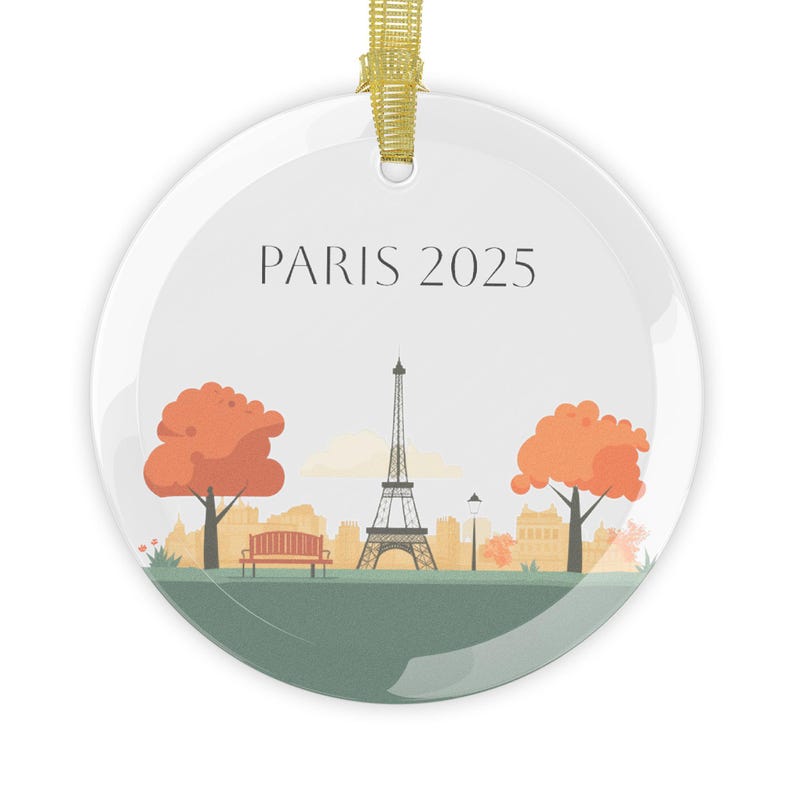 May include: A white round glass ornament with a gold ribbon. The ornament features a stylized illustration of the Eiffel Tower in Paris, France, with two trees on either side of the tower. The text "PARIS 2025" is printed at the top of the ornament.