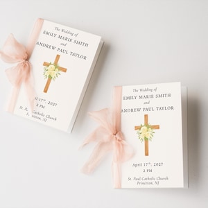 May include: Two white wedding programmes featuring the text The Wedding of Emily Marie Smith and Andrew Paul Taylor, a watercolour cross with white flowers, the date April 17th, 2027, 2 PM, and St. Paul Catholic Church, Princeton, NJ, tied with a sheer peach ribbon.