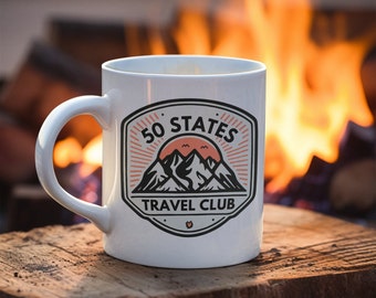 Può includere: Una tazza in ceramica bianca con un logo nero e arancione che dice "50 States Travel Club" e raffigura montagne e un tramonto. La tazza è appoggiata su una superficie di legno davanti a uno sfondo sfocato di un fuoco.