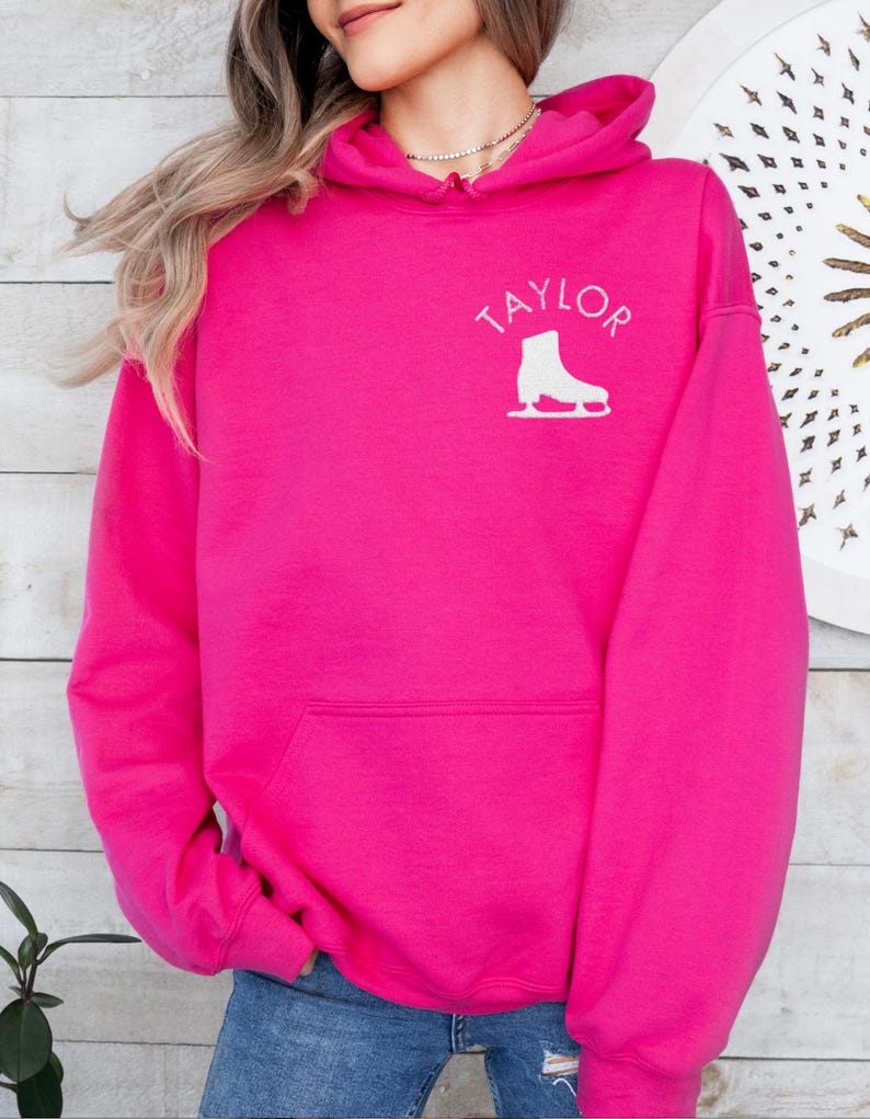 K&ouml;nnte beinhalten: Ein leuchtend pinkfarbener Hoodie mit einer wei&szlig;en Schlittschuhgrafik und dem Namen "TAYLOR" auf der Vorderseite.