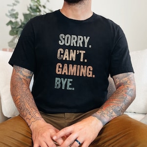 以下が含まれることがあります： 「SORRY. CAN'T. GAMING. BYE.」とヴィンテージスタイルのグラフィックがプリントされた黒いTシャツ