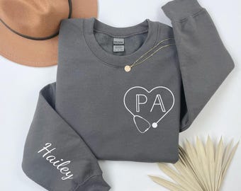 May include: Gray sweatshirt with a white heart design that says "PA" inside the heart. A stethoscope is drawn below the heart. The word "Hailey" is written on the left sleeve.