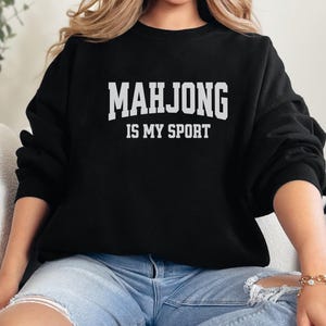 May include: Black sweatshirt featuring the white text "MAHJONG IS MY SPORT". The crew neck sweatshirt is worn with blue jeans. The image presents a relaxed, everyday style, suitable for casual wear.