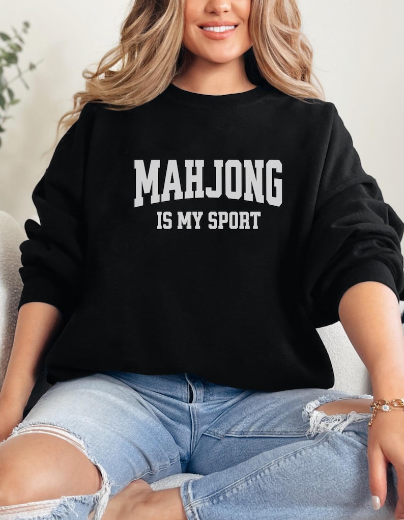 May include: Black sweatshirt featuring the white text "MAHJONG IS MY SPORT". The crew neck sweatshirt is worn with blue jeans. The image presents a relaxed, everyday style, suitable for casual wear.