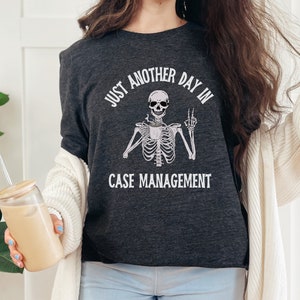 May include: A dark gray t-shirt with a skeleton holding a coffee cup and giving a peace sign. The text on the shirt reads "Just another day in case management."