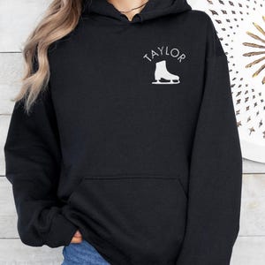 K&ouml;nnte beinhalten: Schwarzer Kapuzenpullover mit einem wei&szlig;en Aufdruck eines Schlittschuhs und dem Wort "TAYLOR" auf der Vorderseite.