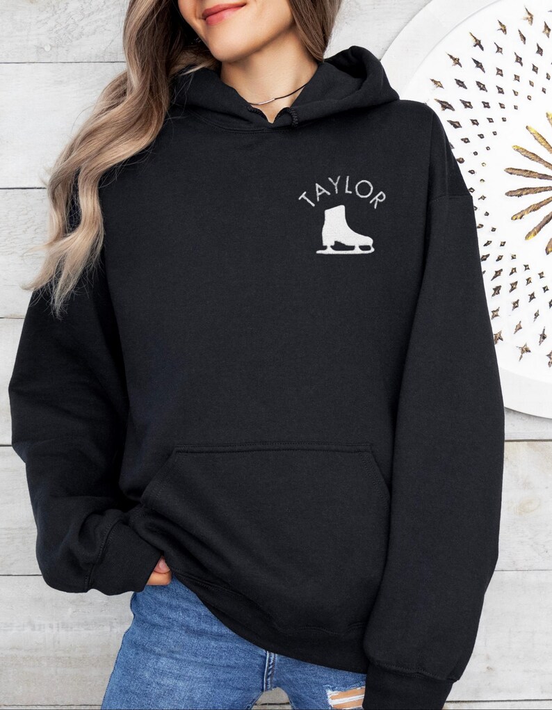 K&ouml;nnte beinhalten: Schwarzer Kapuzenpullover mit einem wei&szlig;en Aufdruck eines Schlittschuhs und dem Wort "TAYLOR" auf der Vorderseite.