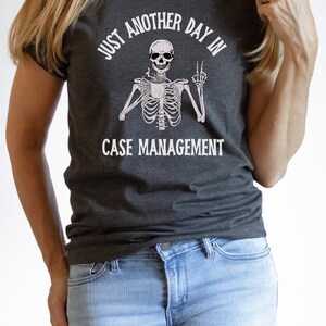 May include: A dark gray t-shirt with a white skeleton graphic holding a coffee cup and making a peace sign. The text "Just another day in case management" is printed on the shirt.