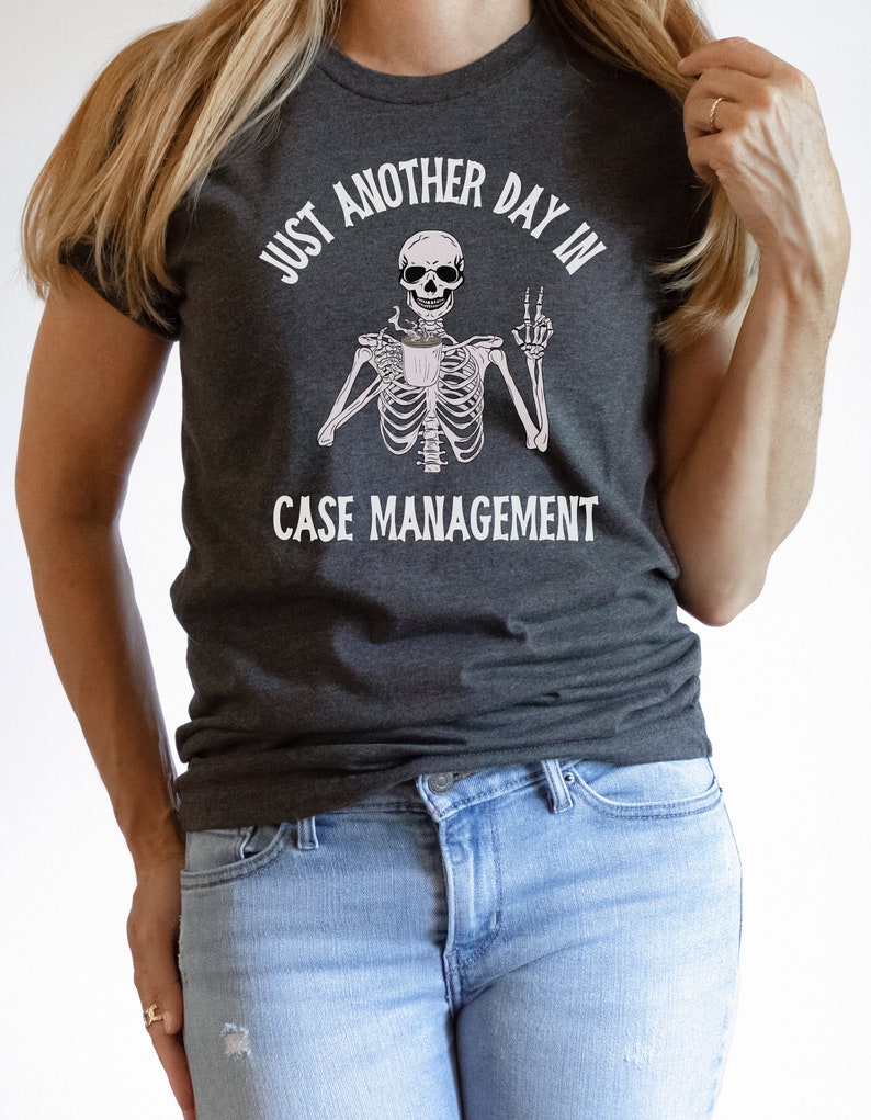 May include: A dark gray t-shirt with a white skeleton graphic holding a coffee cup and making a peace sign. The text "Just another day in case management" is printed on the shirt.