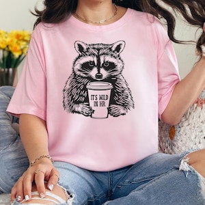 May include: Pale pink t-shirt with a black and white raccoon design. The raccoon holds a coffee cup with the text "IT'S WILD IN HR". The shirt is a relaxed fit and the raccoon is in the center.