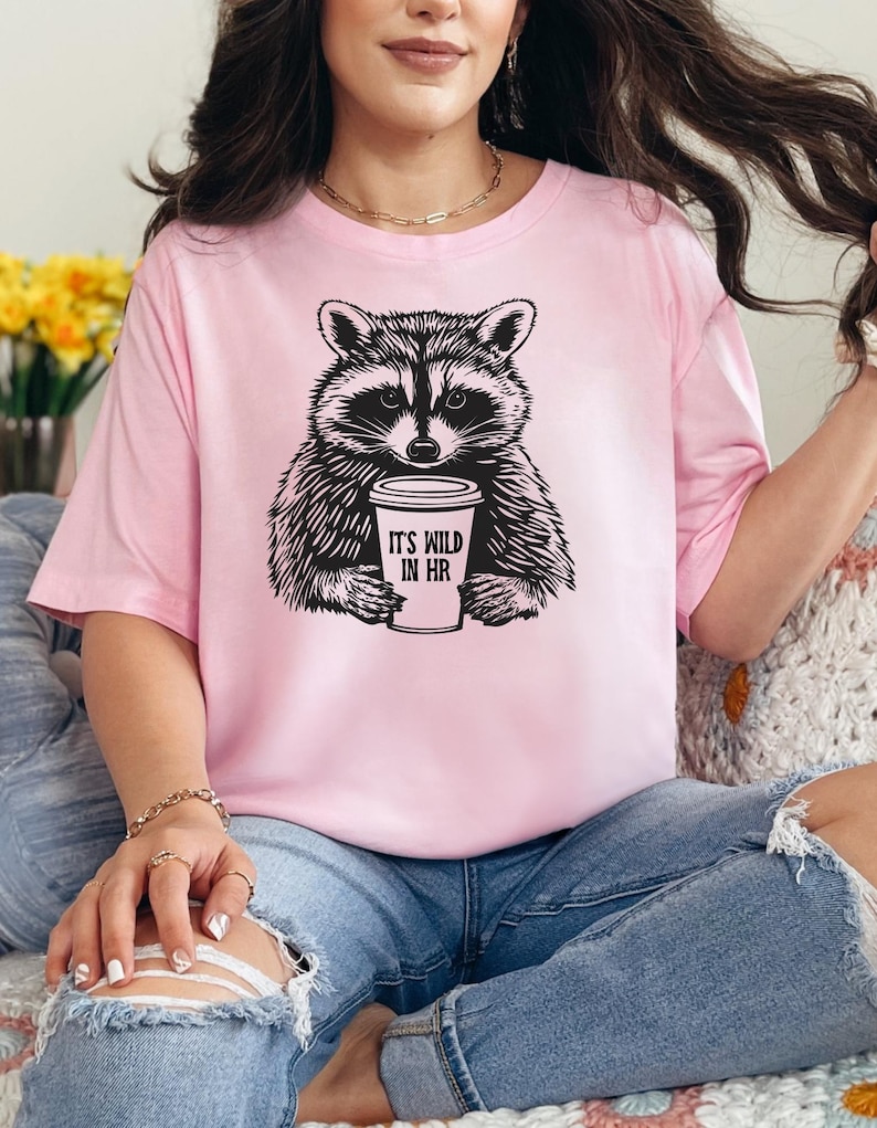 May include: Pale pink t-shirt with a black and white raccoon design. The raccoon holds a coffee cup with the text "IT'S WILD IN HR". The shirt is a relaxed fit and the raccoon is in the center.