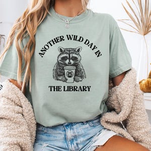 Peut inclure: T-shirt vert sauge avec un raton laveur tenant une tasse de caf&eacute;. Le texte "ANOTHER WILD DAY IN THE LIBRARY" entoure le raton laveur. Le t-shirt est port&eacute; avec un short en jean.