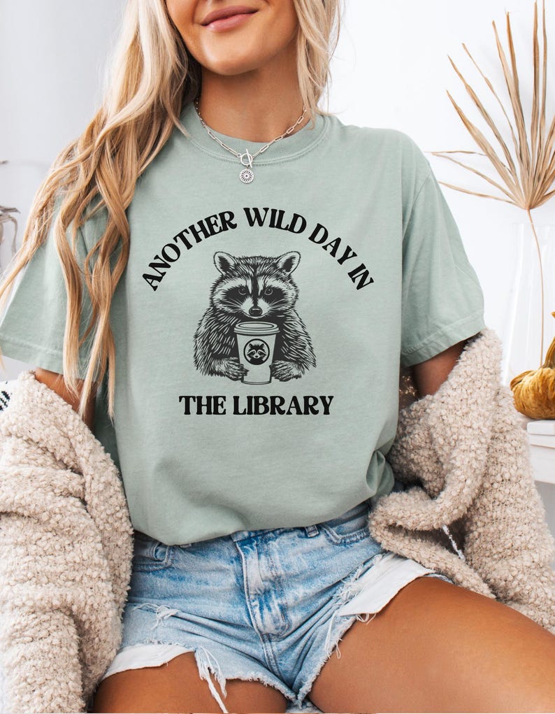 Peut inclure: T-shirt vert sauge avec un raton laveur tenant une tasse de caf&eacute;. Le texte "ANOTHER WILD DAY IN THE LIBRARY" entoure le raton laveur. Le t-shirt est port&eacute; avec un short en jean.