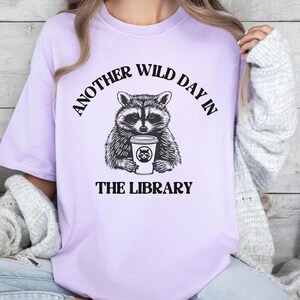 Peut inclure: Un t-shirt lilas clair avec une illustration en noir et blanc d'un raton laveur tenant une tasse de caf&eacute;. Le texte "ANOTHER WILD DAY IN THE LIBRARY" est imprim&eacute; au-dessus et en dessous.