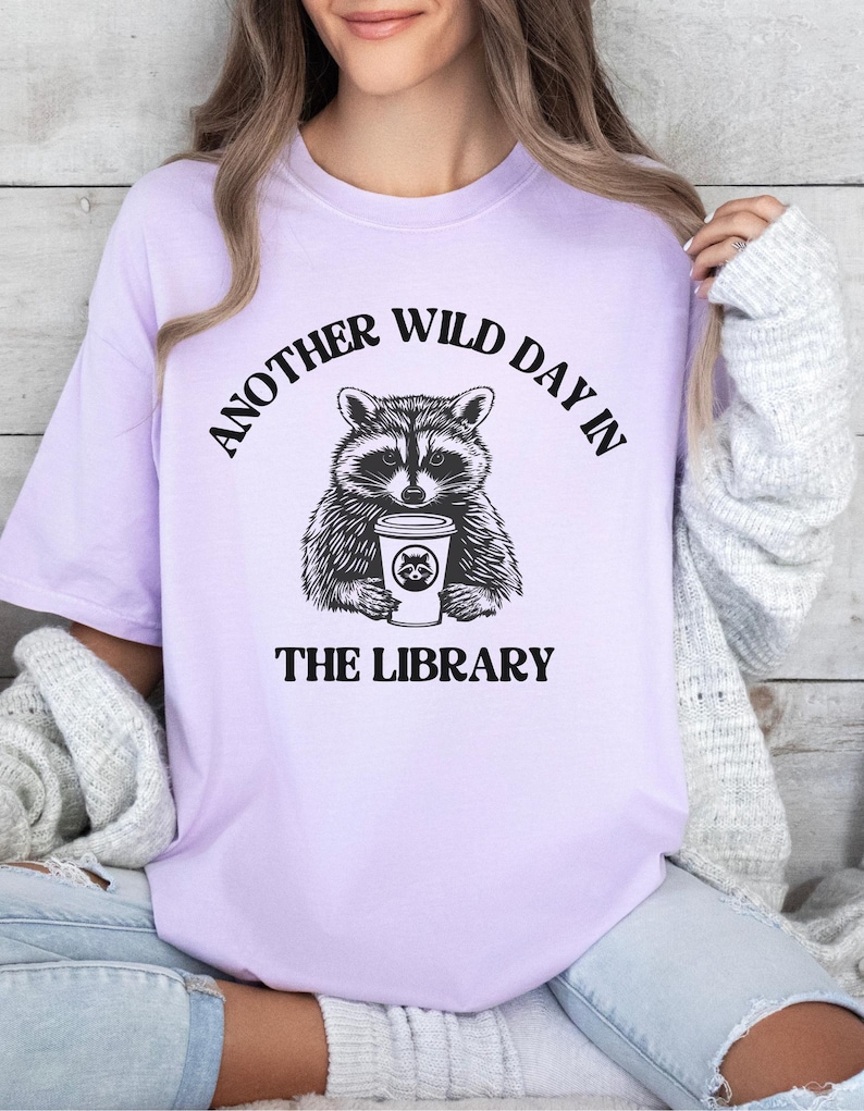 Peut inclure: Un t-shirt lilas clair avec une illustration en noir et blanc d'un raton laveur tenant une tasse de caf&eacute;. Le texte "ANOTHER WILD DAY IN THE LIBRARY" est imprim&eacute; au-dessus et en dessous.