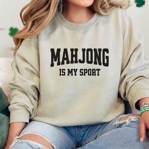 May include: A pale green sweatshirt featuring the black text "MAHJONG IS MY SPORT". The sweatshirt is worn with light blue jeans. The image shows a person smiling.