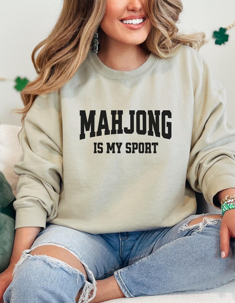 May include: A pale green sweatshirt featuring the black text "MAHJONG IS MY SPORT". The sweatshirt is worn with light blue jeans. The image shows a person smiling.