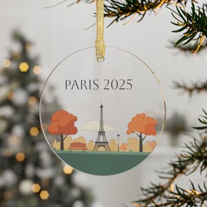 May include: A round, clear glass ornament with a gold ribbon. The ornament features a stylized illustration of the Eiffel Tower in Paris, France, with two trees on either side. The text "PARIS 2025" is printed at the top of the ornament.