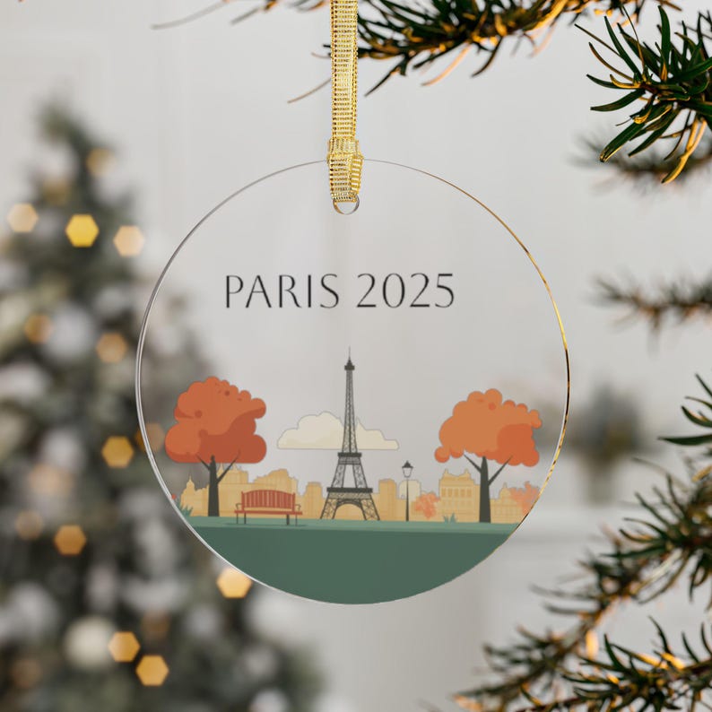May include: A round, clear glass ornament with a gold ribbon. The ornament features a stylized illustration of the Eiffel Tower in Paris, France, with two trees on either side. The text "PARIS 2025" is printed at the top of the ornament.