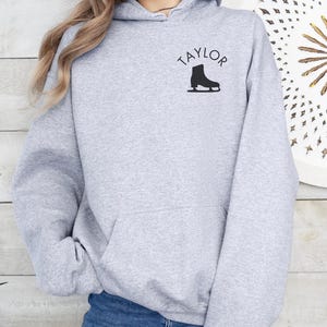 K&ouml;nnte beinhalten: Grauer Kapuzenpullover mit dem Wort "TAYLOR" und einer Silhouette einer Schlittschuhs auf der Vorderseite.