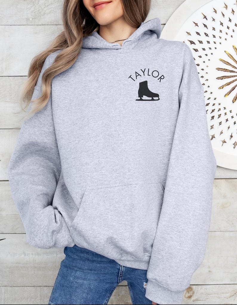 K&ouml;nnte beinhalten: Grauer Kapuzenpullover mit dem Wort "TAYLOR" und einer Silhouette einer Schlittschuhs auf der Vorderseite.