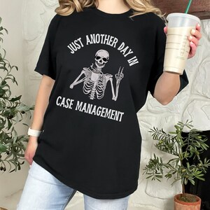 May include: Black t-shirt with a white skeleton graphic holding a coffee cup and the text "Just another day in case management".