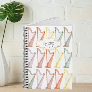 Op de afbeelding: Een spiraalgebonden notitieboek met een witte omslag met een herhalend patroon van aquarel-achtige harpen in verschillende pastelkleuren. De omslag heeft het woord "Notes" in zwart gedrukt op een gouden rechthoek.