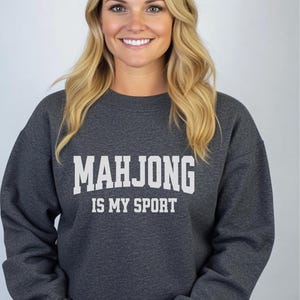 May include: A dark grey crew neck sweatshirt with the white text "MAHJONG IS MY SPORT". The sweatshirt is made of a soft material. The image is of a person wearing the sweatshirt. The theme is related to games and hobbies.