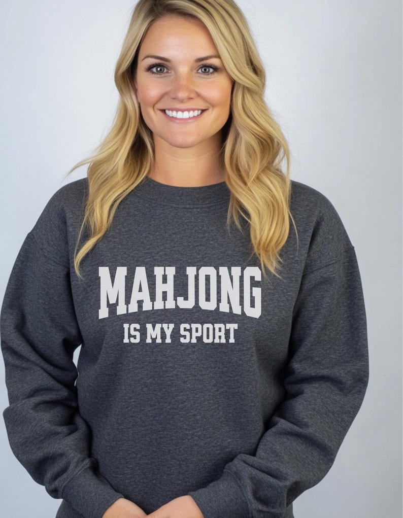 May include: A dark grey crew neck sweatshirt with the white text "MAHJONG IS MY SPORT". The sweatshirt is made of a soft material. The image is of a person wearing the sweatshirt. The theme is related to games and hobbies.