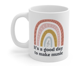 Może przedstawiać: Biały kubek ceramiczny z wzorem tęczy z nutami muzycznymi i sercami. Pod tęczą nadrukowany jest tekst "It's a good day to make music".